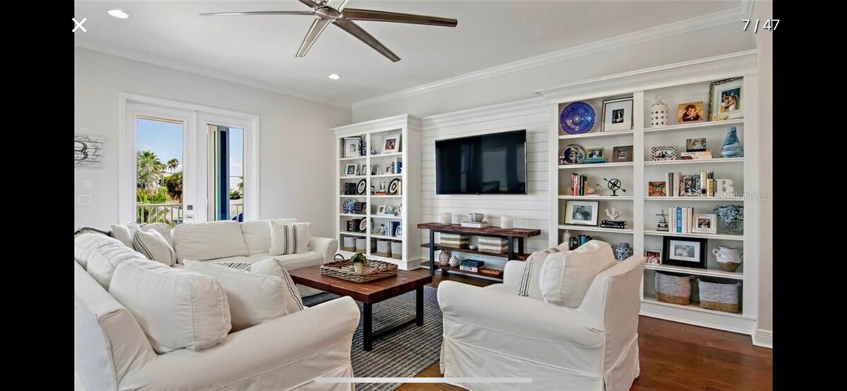 A modern living room features a large flat screen TV mounted on a white wall, flanked by built-in shelves showcasing decorative items and personal photos. Comfortable white sofas are arranged around a dark wood coffee table, creating an inviting atmosphere with natural light flowing through double doors.