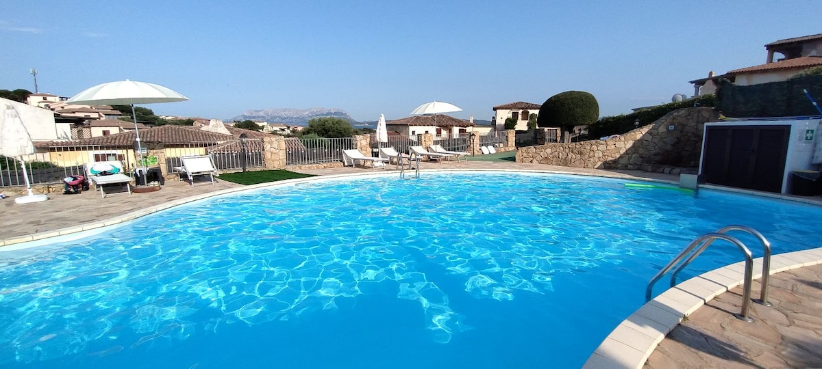 A shared swimming pool shimmers under the clear sky, surrounded by sun loungers and umbrellas. A stone perimeter encloses the area, enhancing the tranquil outdoor setting. Views of the surrounding landscape and distant hills complete the inviting atmosphere for relaxation and leisure.