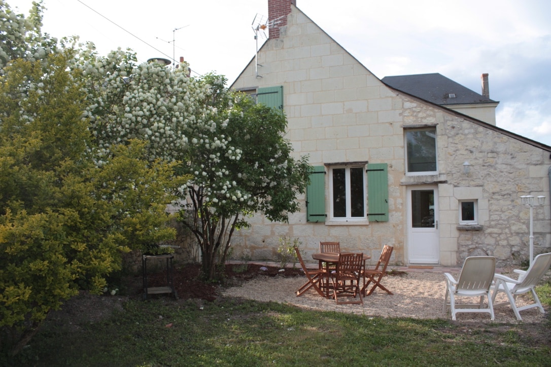 The exterior features a charming stone house with green shutters, surrounded by a garden with flowering shrubs. A gravel area includes a wooden dining table and chairs complemented by white lounge chairs, creating a welcoming space for outdoor relaxation.
