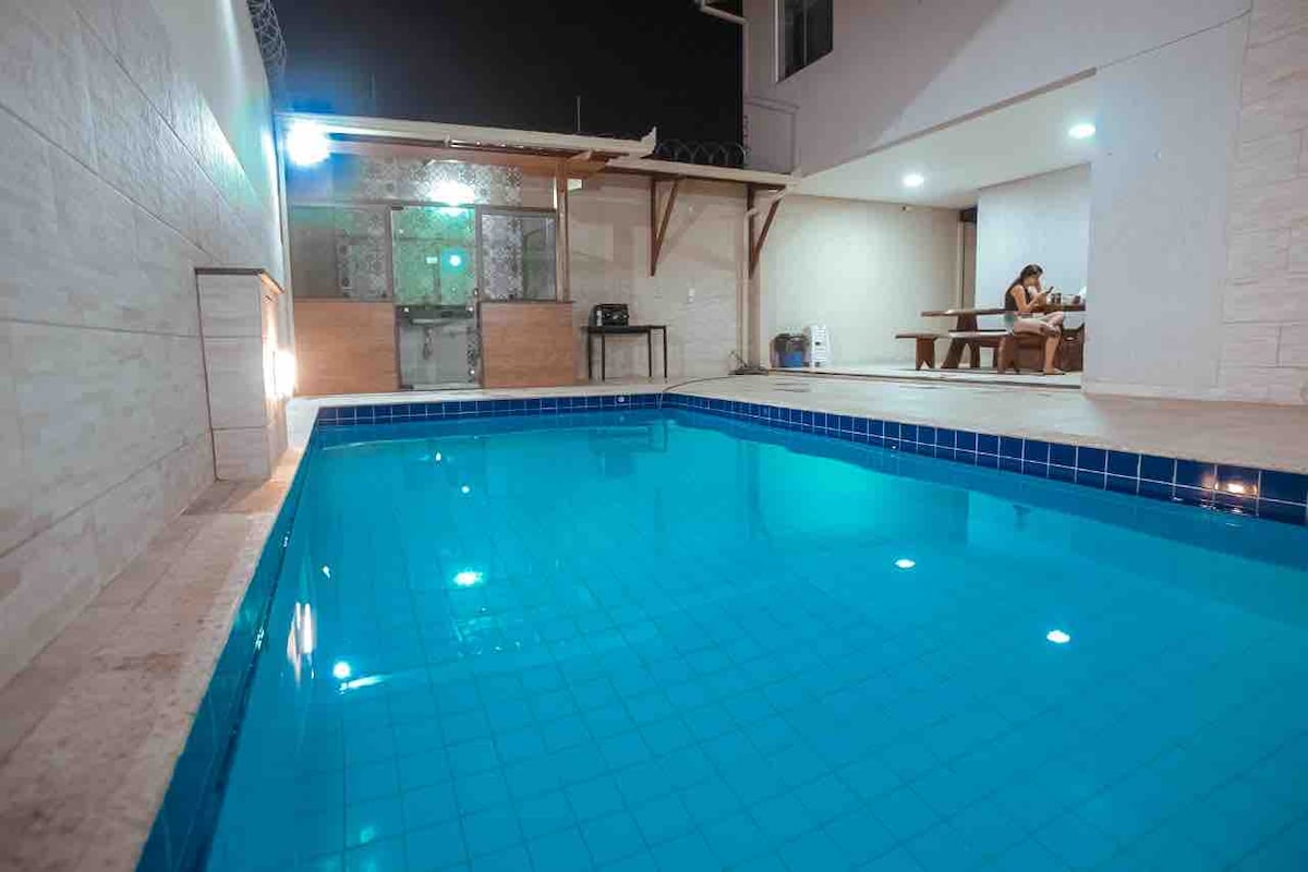 A tranquil swimming pool featuring clear blue water reflects ambient lighting, contributing to a serene environment. A covered seating area with a table is visible in the background, suggesting a relaxing space for socializing or dining.