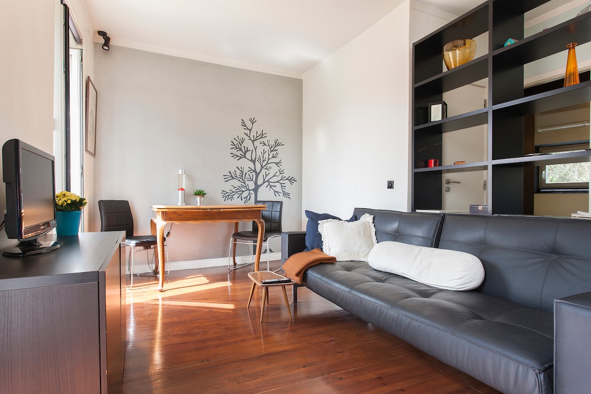 A bright living room features a dark leather sofa, complemented by a soft white pillow. A wooden dining table is positioned nearby, accompanied by black chairs. Natural light streams through large windows, illuminating the space adorned with a decorative tree wall art.