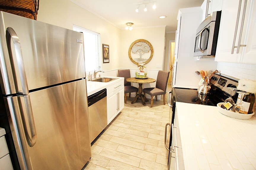 A modern kitchen features stainless steel appliances, including a refrigerator, dishwasher, and oven. Light-colored cabinets and tile backsplash complement the bright countertops. A small round table is set with two upholstered chairs, while natural light streams through the window, enhancing the space's functionality.