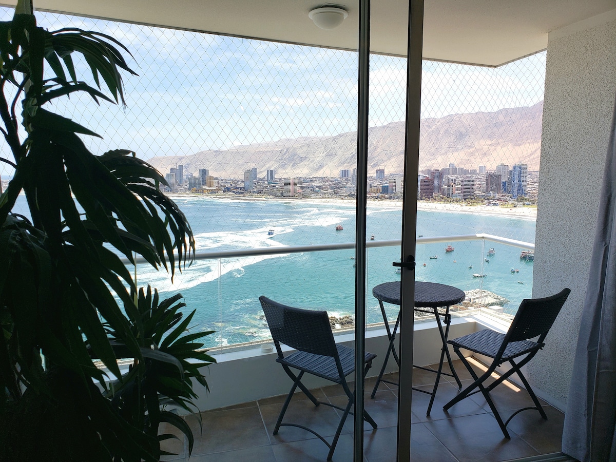 A balcony area features a small round table and two chairs, offering views of the ocean and surrounding cityscape. The scene is framed by a green plant, with mesh safety netting providing a sense of security while enjoying the scenery.