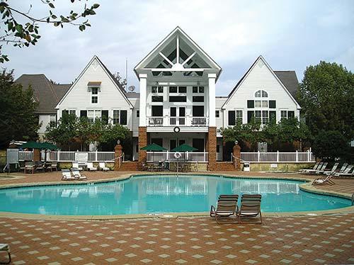 A large outdoor pool is framed by a spacious deck with lounge chairs, leading to a three-story white building with a peaked roof. Trees provide shade, and the area is well-maintained, creating an inviting recreational space.