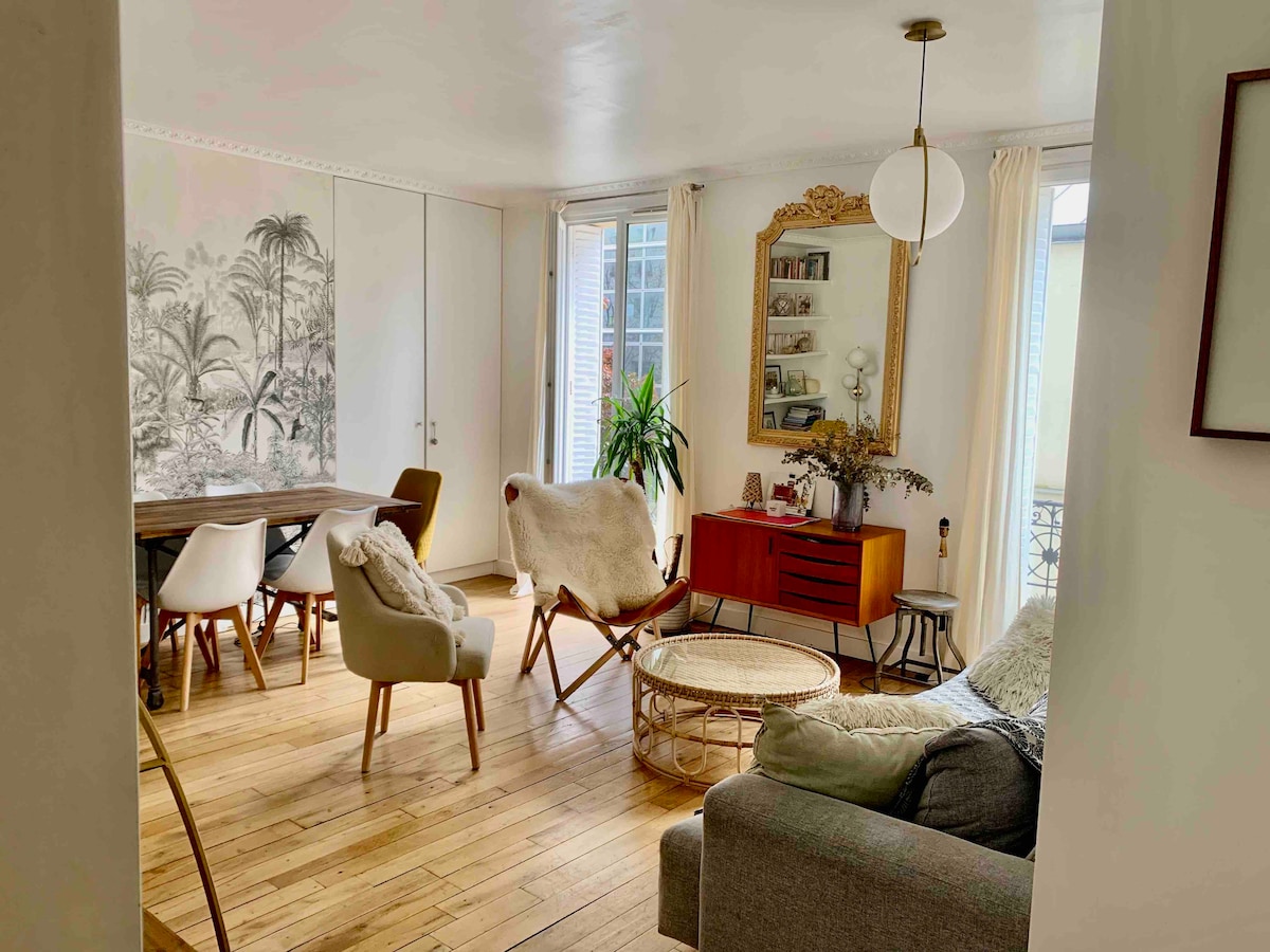 A sunny living room features wooden floors and large windows allowing natural light. A mix of seating options includes a cozy sofa, armchairs, and a round coffee table. Decorative elements such as a plant and a chic mirror add character to the space.