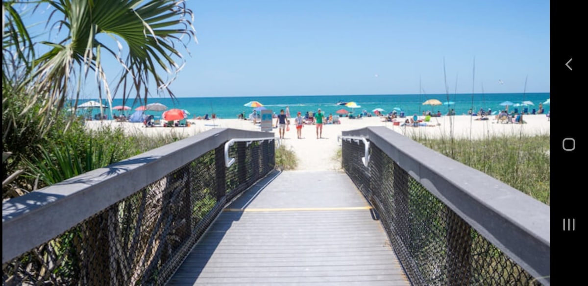 A wooden boardwalk leads directly to a sandy beach, framed by lush greenery on either side. Sunbathers relax under colorful umbrellas, with the calm turquoise waters visible in the background. The scene captures a sunny day, inviting relaxation and enjoyment.