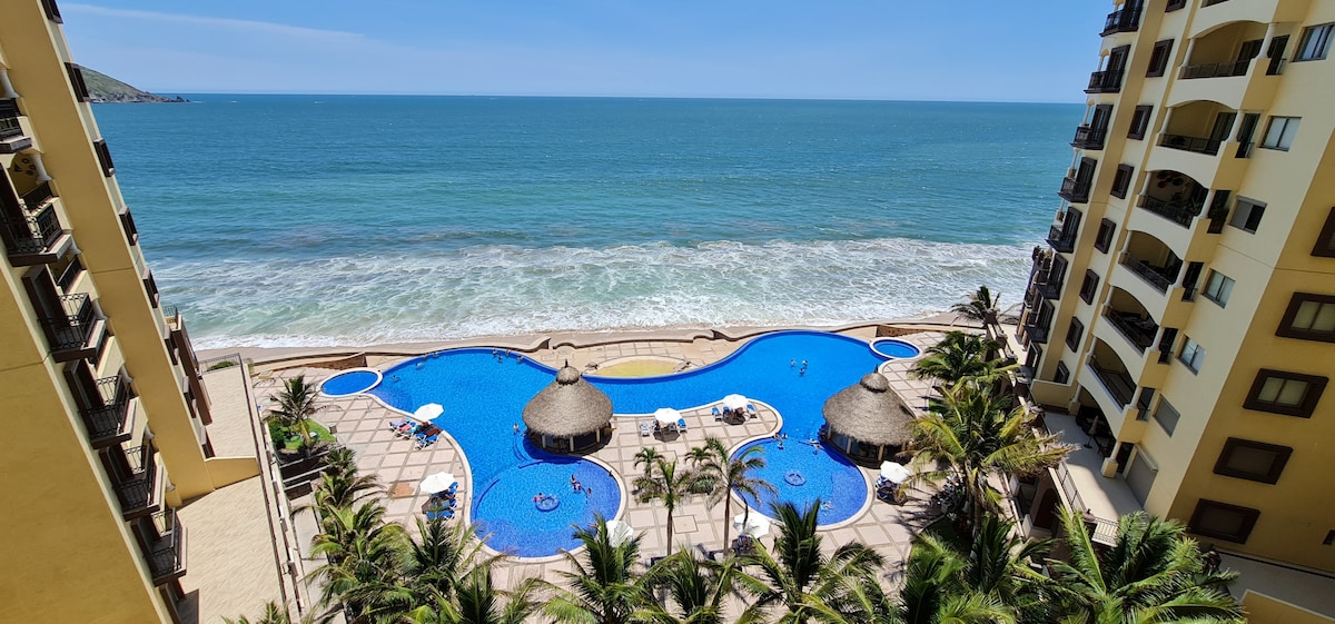 An infinity pool is framed by palm trees and features thatched-roof umbrellas scattered around its edge. The bright blue water contrasts with the vibrant ocean waves visible in the background, highlighting the spacious outdoor area and the scenic coastline.