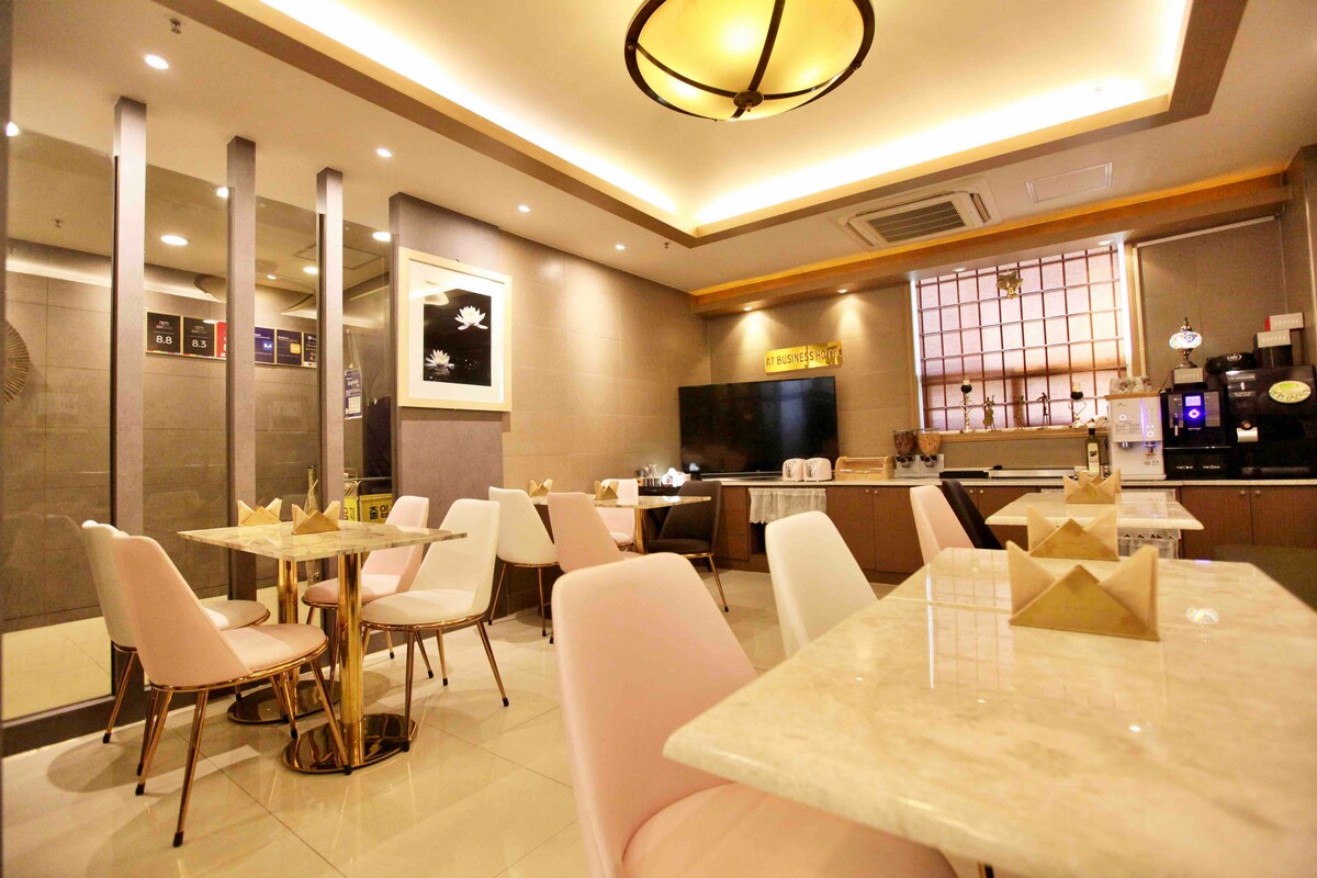 A modern dining area is equipped with sleek marble tables and soft pink chairs, arranged for a pleasant ambiance. A coffee station is visible against a backdrop of framed artwork and warm lighting, creating an inviting space for guests to enjoy their meals.