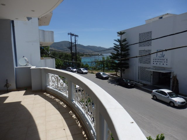 A spacious balcony is visible, featuring ornate railings that overlook the street and the sea beyond. Vehicles are parked along the road, with trees lining the background against a clear blue sky. The scene is tranquil, highlighting the proximity to the waterfront.