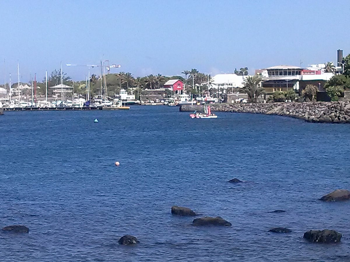 A view of a calm harbor is captured, featuring several moored boats and gentle waves reflecting sunlight. On the shore, buildings are visible among palm trees, with a vibrant red structure adding a pop of color to the landscape. Large rocks border the water's edge.