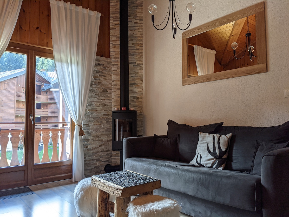 A cozy sitting area features a comfortable sofa with decorative pillows, positioned near large glass doors that lead to a balcony. A wooden coffee table rests on a light floor, and a decorative chandelier adds a modern touch. Natural light fills the space.
