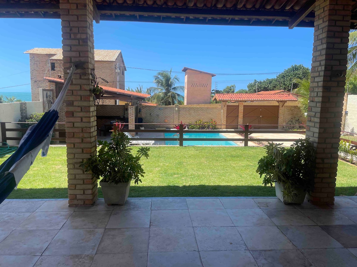 A covered patio area features a hammock and potted plants, providing a view of the well-maintained lawn and pool area. The background includes a brick structure and palm trees, with clear skies above, suggesting a relaxed outdoor environment.