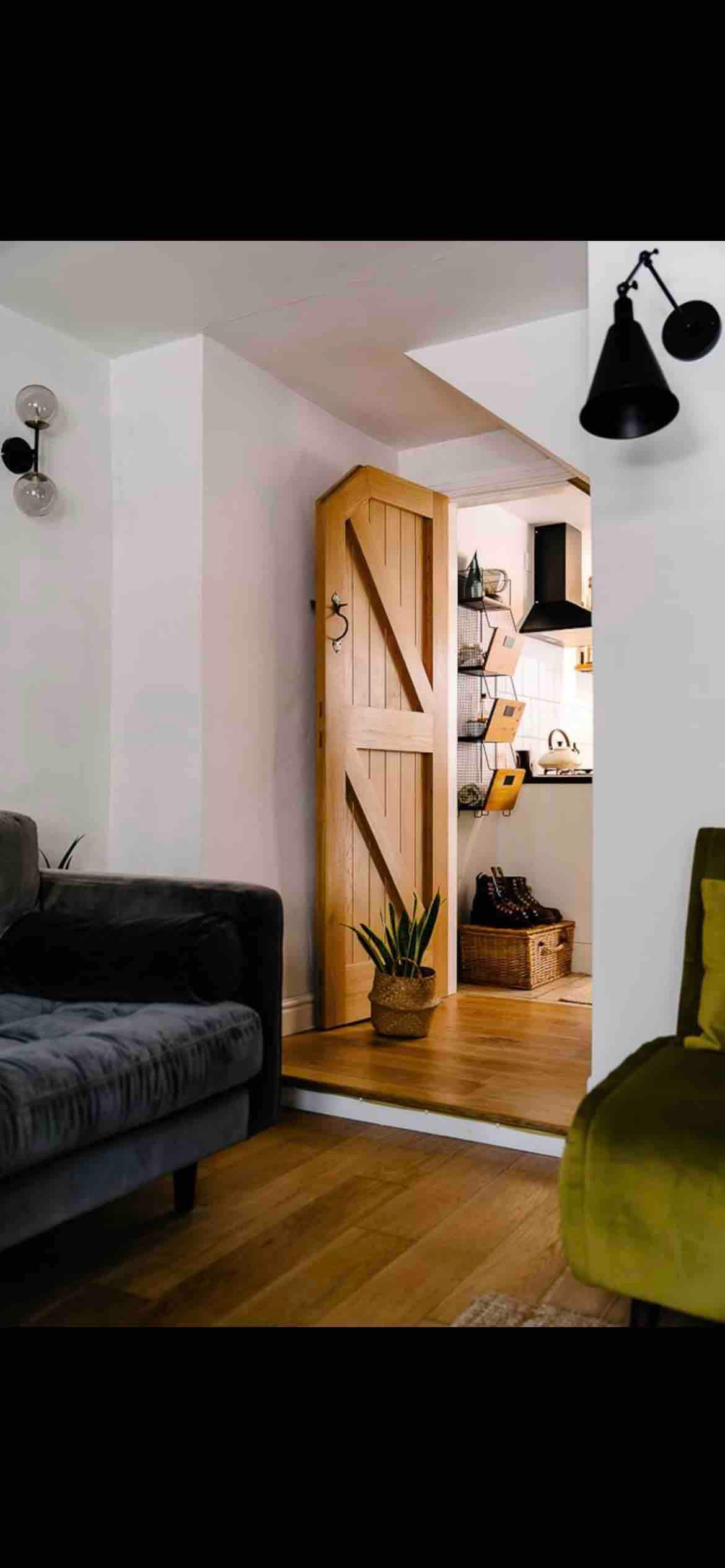 A cozy lounge area features a plush grey sofa and a vibrant green accent chair. A wooden door is visible in the background, leading to a well-lit kitchen area. Natural light enhances the inviting interior, complemented by stylish wall lights.
