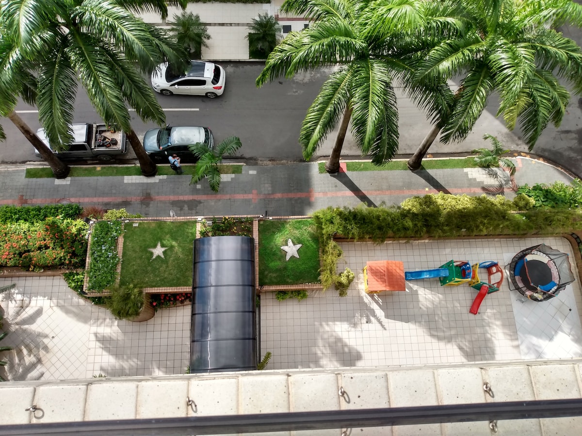 An aerial view showcases a vibrant outdoor area featuring lush greenery and palm trees. A play structure lies beside a trampoline, with geometric designs visible in the landscaping. Paved walkways meander through the space, providing access to the building's main entrance.