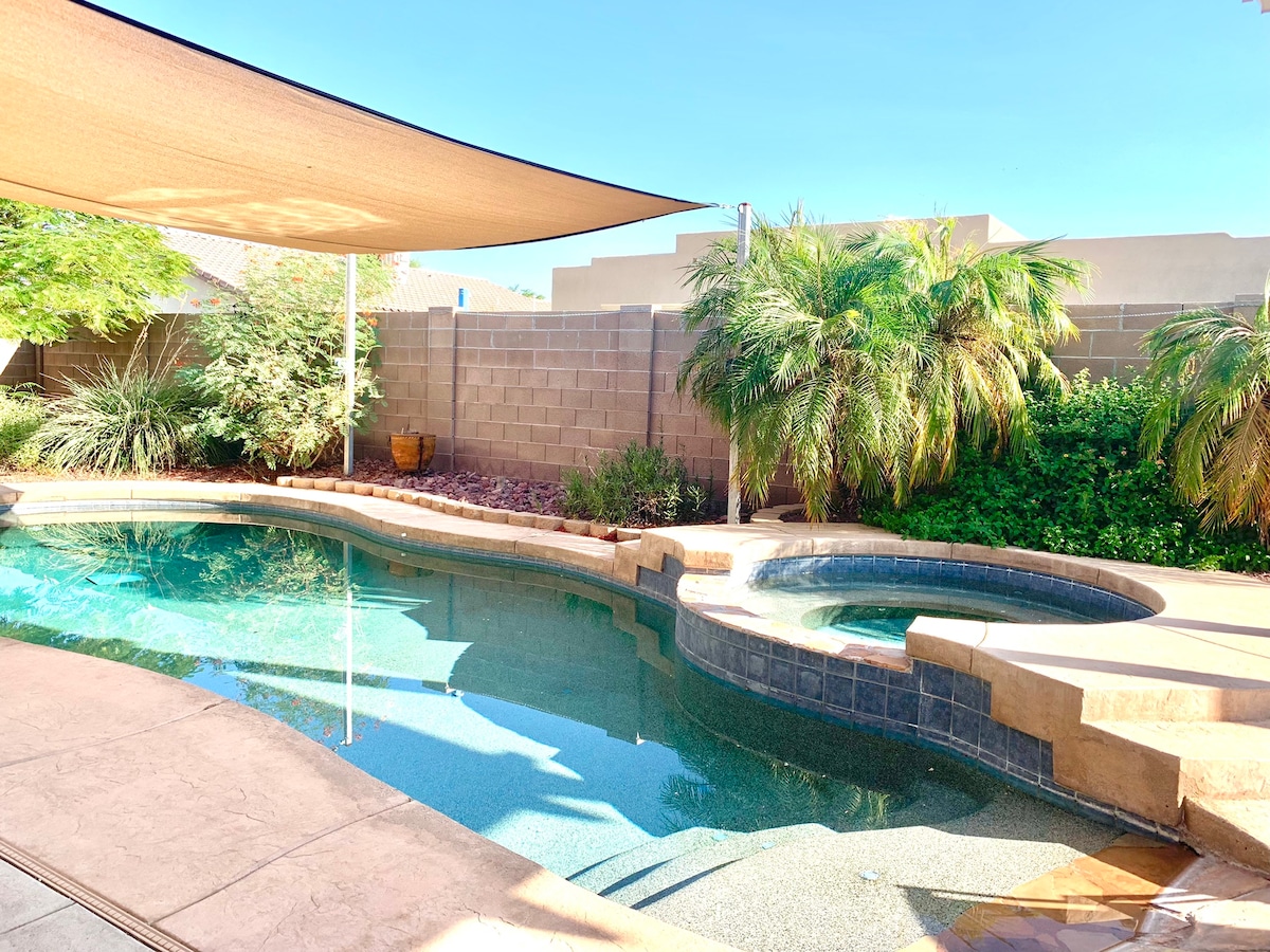 A serene backyard oasis features a salt-water pool surrounded by lush native plants and palm trees. A partially shaded area offers relief from the sun, creating a welcoming space for relaxation. The pool's gentle curves lead to a built-in spa for added comfort.