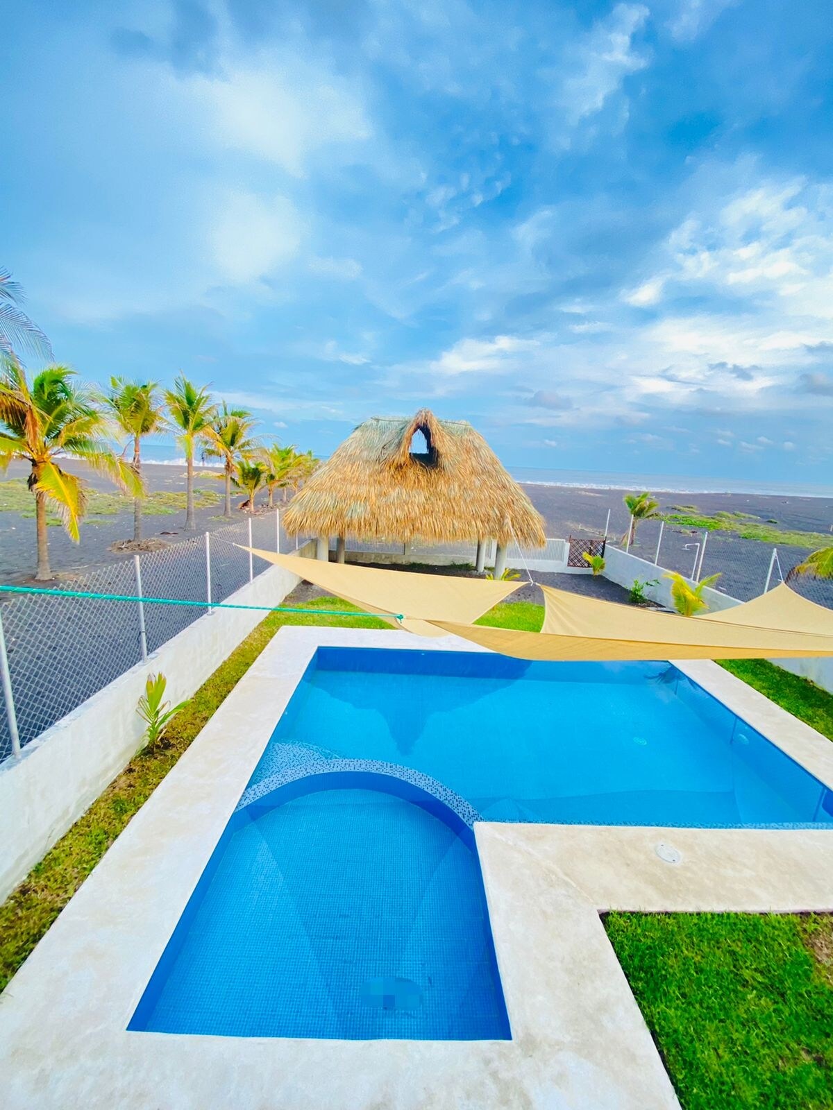 A spacious pool area is featured, surrounded by lush greenery and palm trees. A thatched-roof rancho is visible in the background, and sunshade structures provide comfort. The pool design includes a shallow section for children, and the perimeter is secured with a low fence.