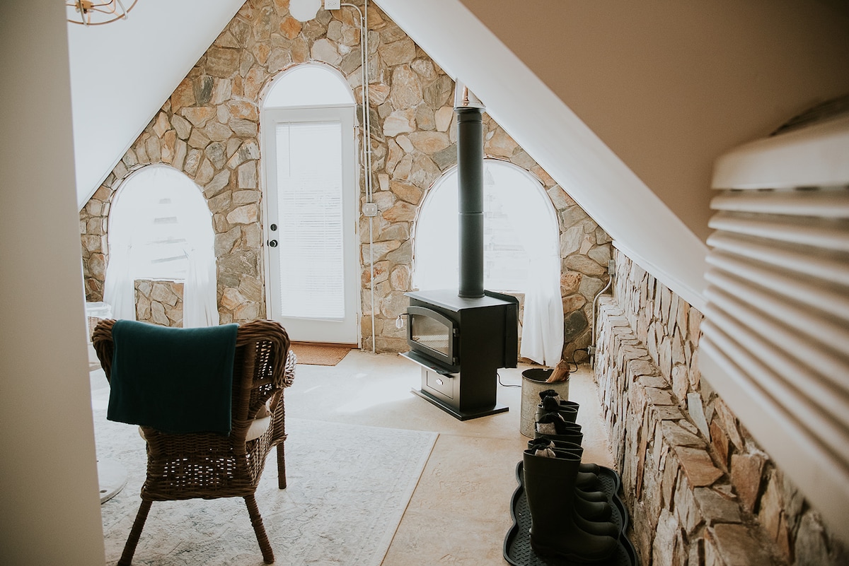 The spacious room features a stone accent wall with arched windows allowing natural light. A wood-burning stove is situated beside a cozy seating area with a wicker chair. The muted tones and textures create an inviting space for relaxation.