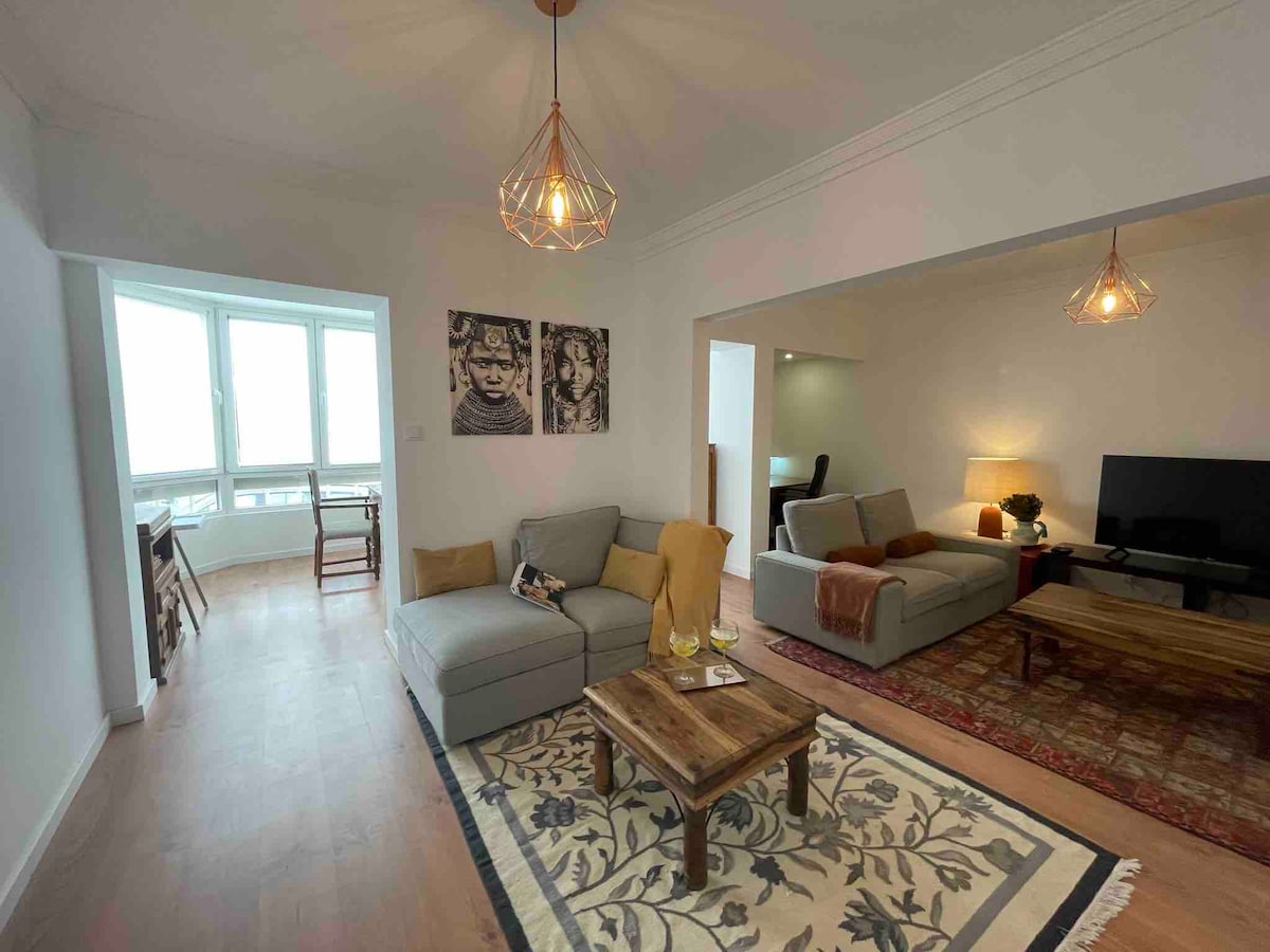 A bright and spacious living area features a light grey sofa set adorned with a cozy throw and decorative cushions. A wooden coffee table sits on an ornate area rug, complemented by modern lighting fixtures. Large windows allow natural light to fill the space, enhancing the inviting atmosphere.