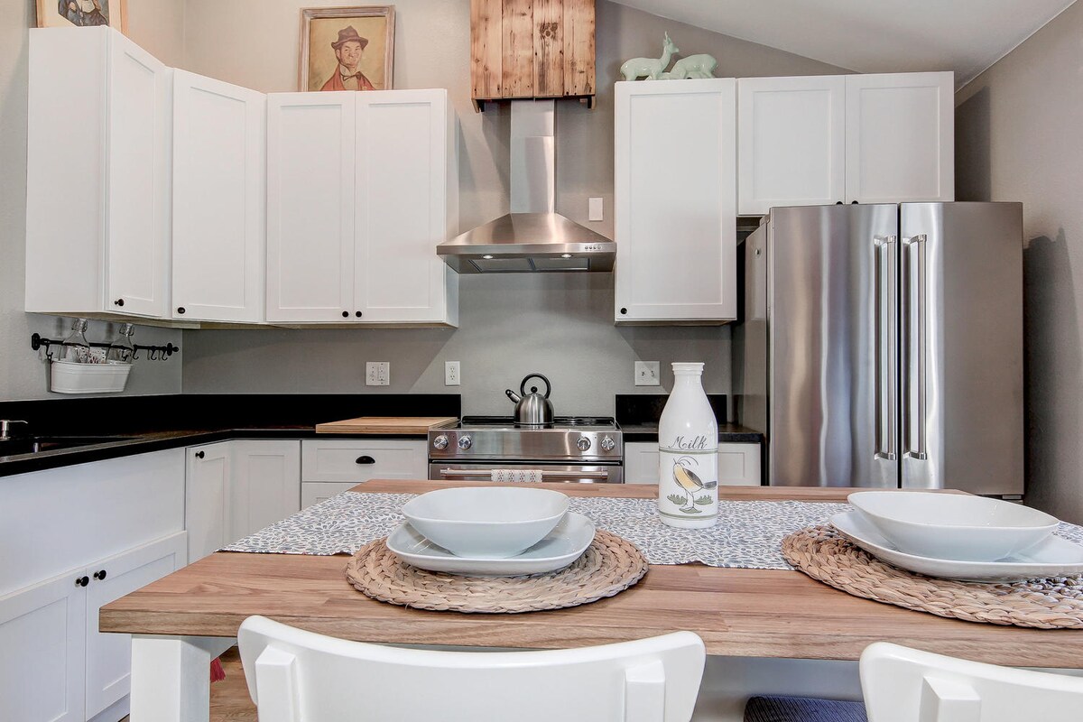 A modern kitchen is presented with white cabinetry and sleek stainless-steel appliances. A wooden dining table is set with two placemats and bowls, inviting a shared meal. Natural light enhances the open layout, while decorative accents add a personal touch.