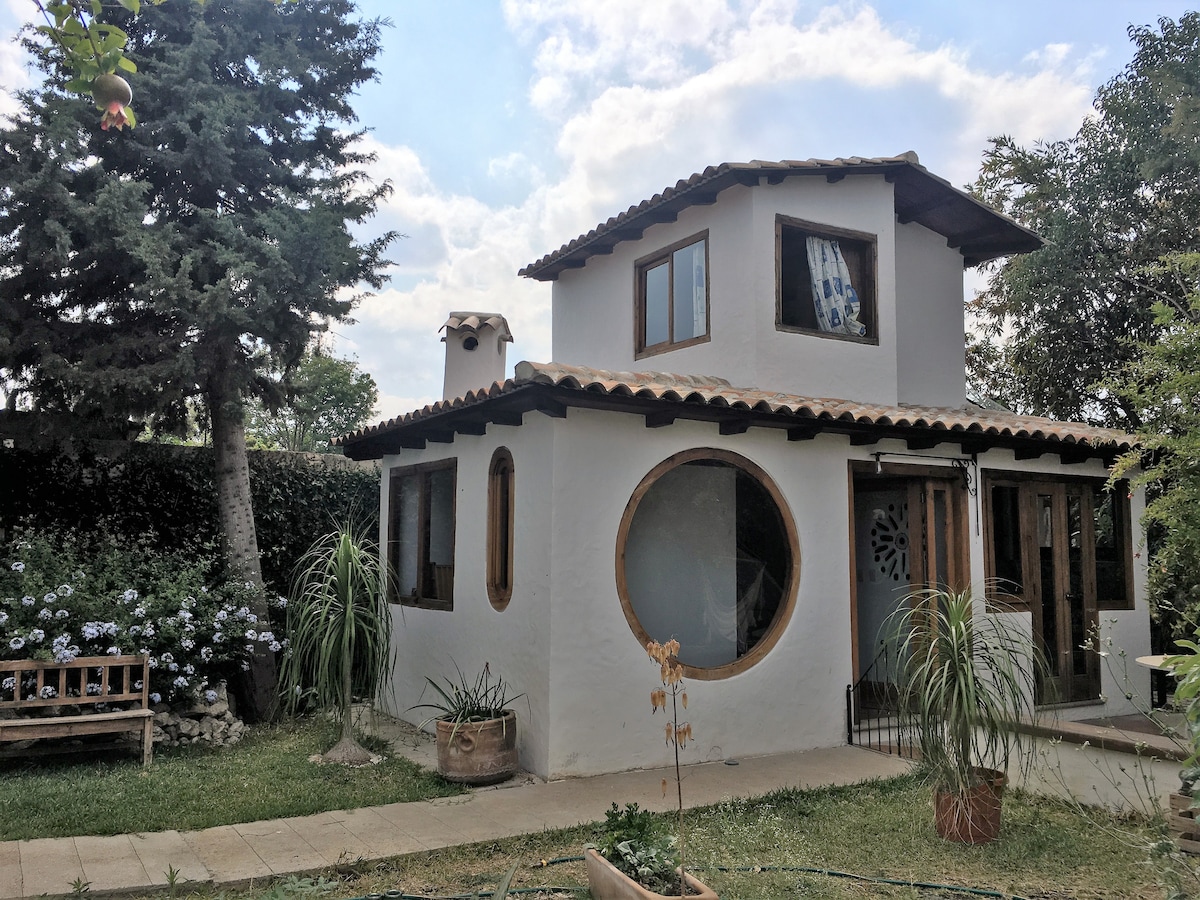 A charming two-story house is set within a lush garden. Large windows complement the circular design feature, allowing natural light to fill the space. A cozy bench is positioned near the entrance, surrounded by vibrant greenery and flowering plants.