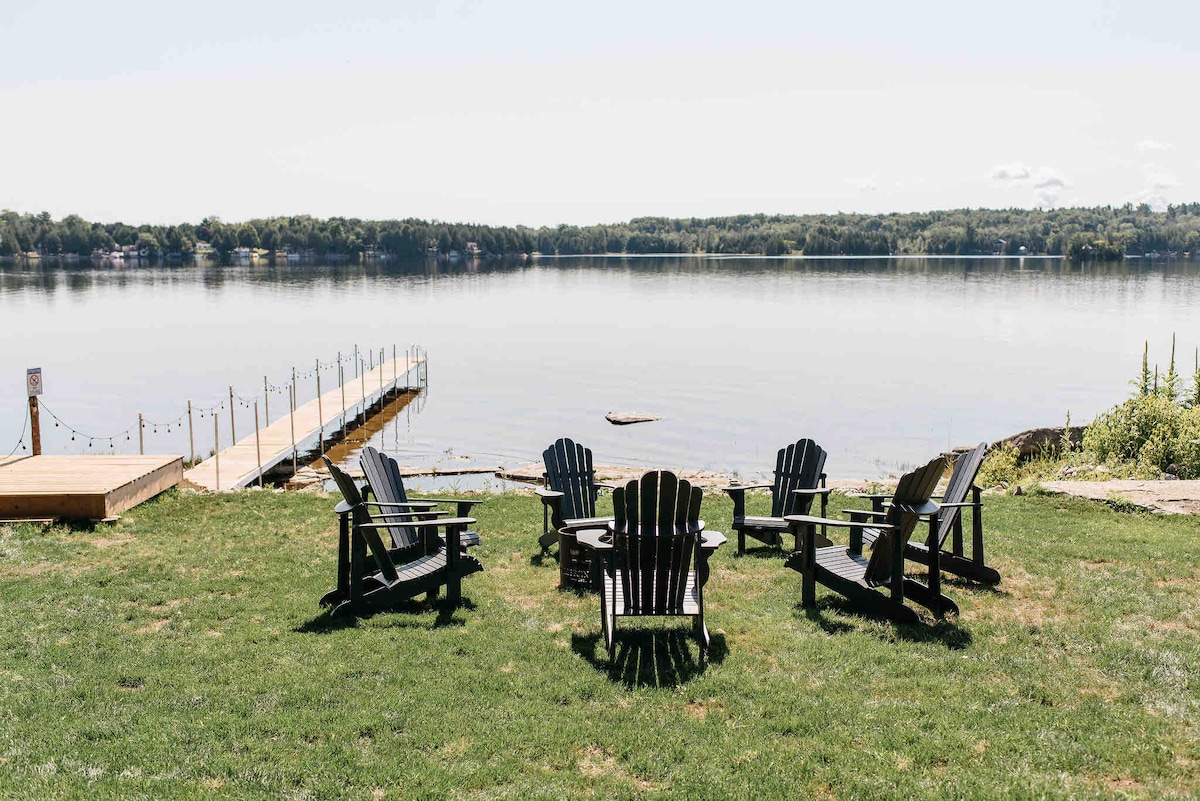 A serene outdoor space features a circle of Adirondack chairs surrounding a central firepit, facing a calm lake. In the background, a wooden dock extends into the water, with gentle waves lapping against the shore. The lush green lawn completes the peaceful scene.