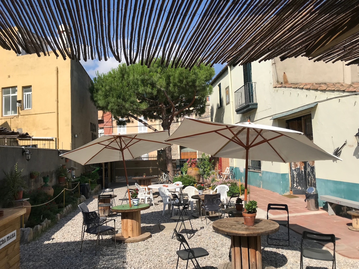 A spacious outdoor area is equipped with large umbrellas providing shade over several tables and chairs. Potted plants add greenery to the gravel-covered ground, while a backdrop of colorful buildings and trees enhances the inviting atmosphere.