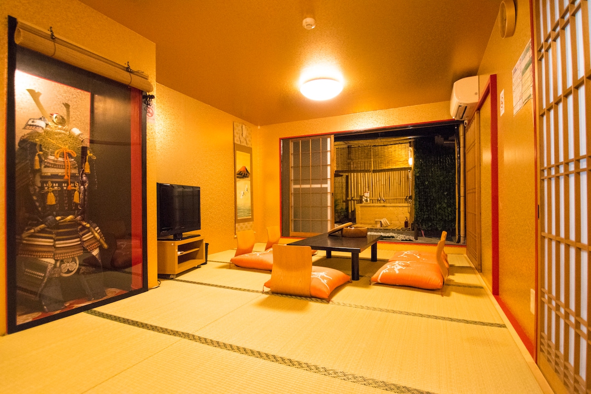 A spacious traditional Japanese sitting room features tatami mat flooring and bright orange walls. A low wooden table is surrounded by cushioned floor seating. A television is positioned against one wall, while sliding doors lead to an outdoor area, allowing natural light to fill the space.