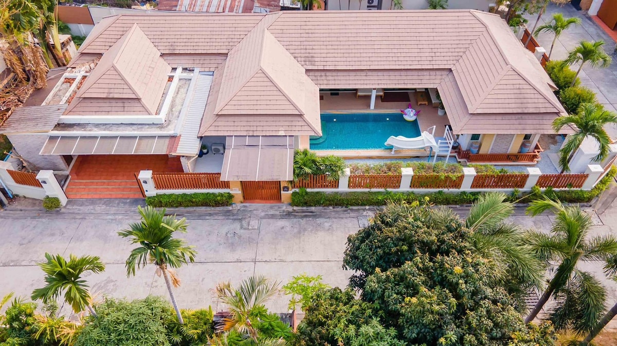 An aerial view of a large villa showcases a spacious layout, featuring a private swimming pool surrounded by palm trees. The building is designed with a triangular roof and is complemented by a well-maintained garden and driveway.