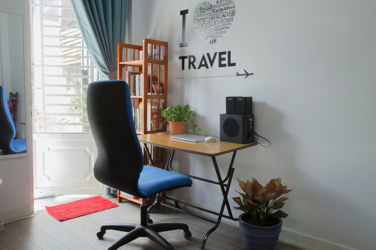 A functional workspace is provided, featuring a wooden desk with a blue office chair. A speaker system and potted plants are positioned on the desk, while a bookshelf stands nearby. Natural light enters through the window, complemented by a red mat on the floor.