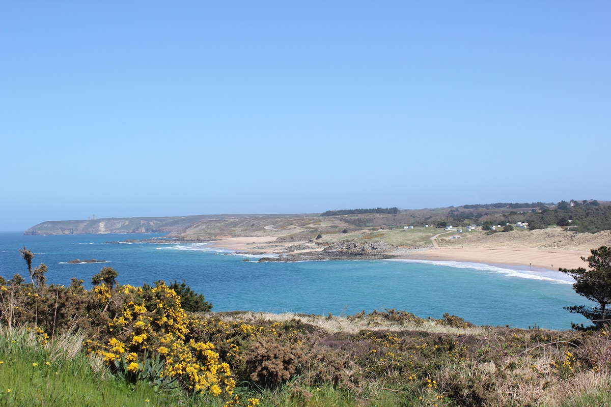 The image shows a serene coastal landscape with a sandy beach stretching along the shoreline. Sparkling blue waters gently lap against the shore, contrasting with the greenery in the foreground. The hills in the distance are dotted with trees, creating a tranquil natural setting.