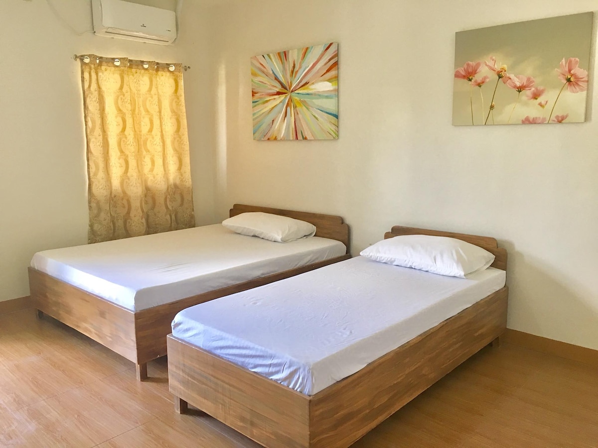 A bright room features two wooden beds with clean white linens, providing a minimalist look. Light natural wood flooring complements the space. Colorful wall art adorns the walls, while a window dressed with sheer curtains allows soft light to filter into the room.