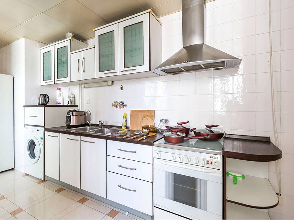 A modern kitchen is equipped with white cabinets and a stainless steel refrigerator. Cooking appliances include an oven and a range hood, with pots arranged on the countertop. A washing machine is situated near the fridge, contributing to the kitchen's functionality.