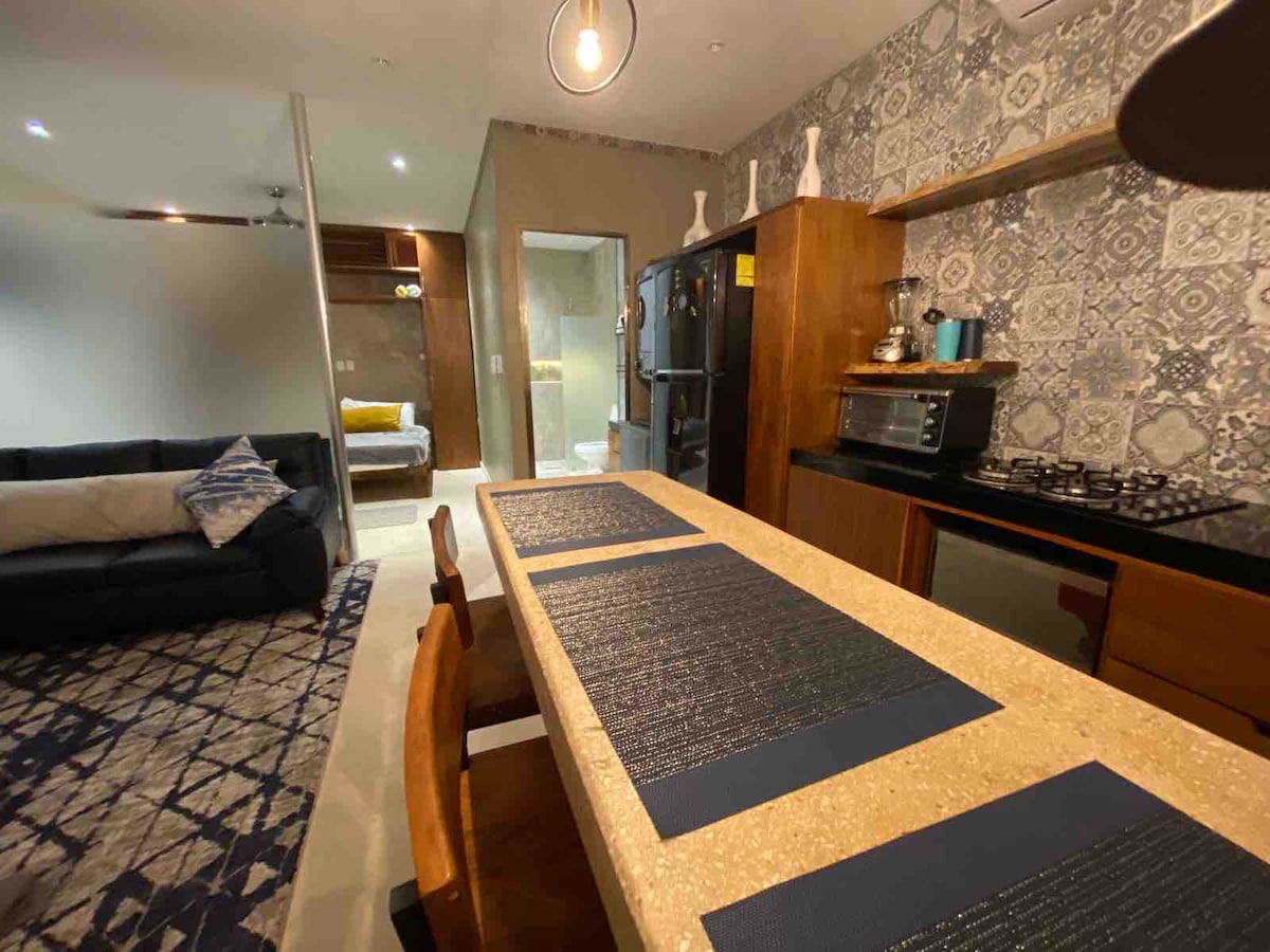 An open kitchen features a stylish countertop with four chairs, complemented by wooden cabinetry and a refrigerator. The adjacent lounge area displays a modern sofa in a neutral tone. A well-designed wall showcases patterned tiles, adding a contemporary touch to the space.