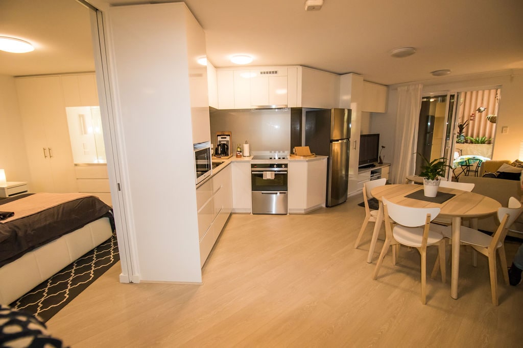 The open-plan living area features a modern kitchen with sleek cabinetry and stainless steel appliances, including a refrigerator and oven. A dining table and chairs are included, while a separate bedroom is visible in the background, enhancing the apartment's spacious layout.
