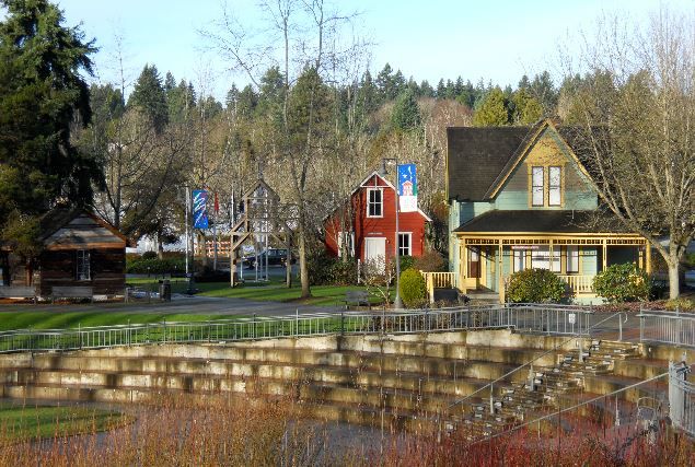 A park setting features a stone terrace leading down to a landscaped area. Distinctive houses with various colors, including red and blue, are visible in the background, surrounded by greenery and trees. Clear skies provide bright lighting to the scene.