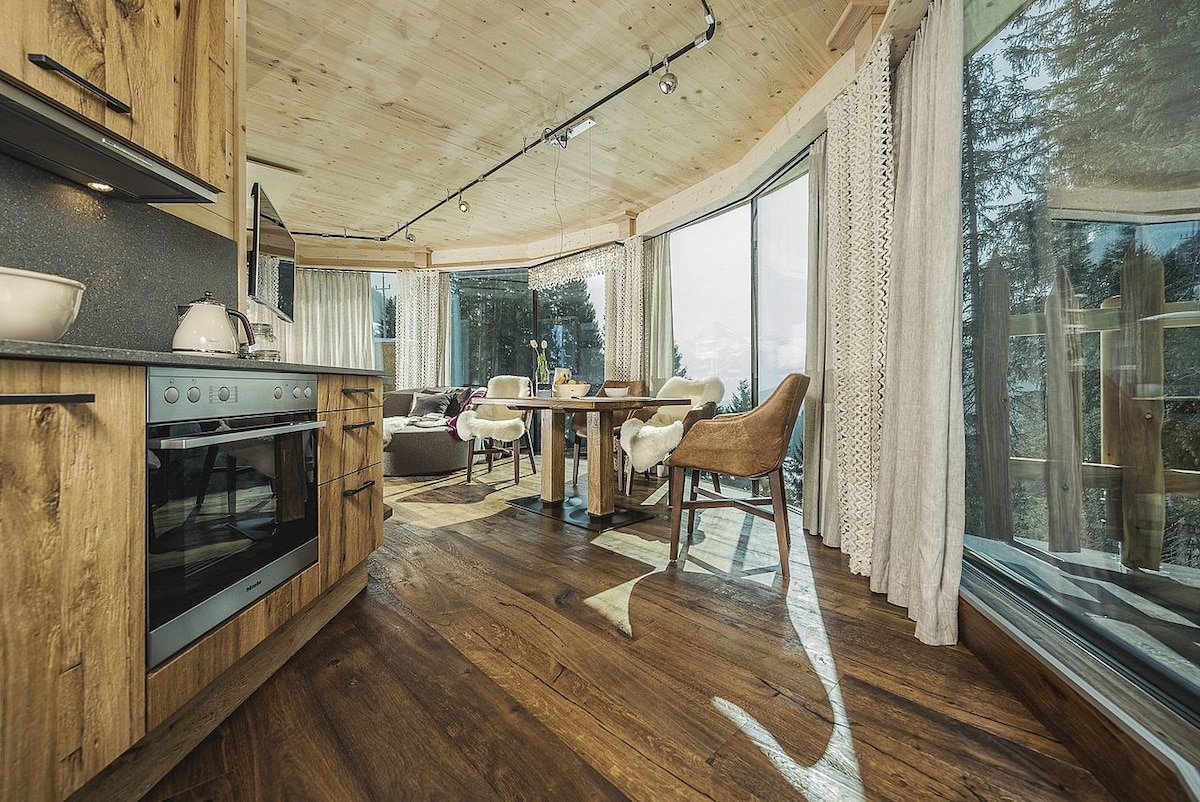 A bright dining area features large windows that invite natural light, highlighting a wooden table surrounded by comfortable chairs. The kitchen area, with sleek cabinets, complements the open design, creating an inviting space for meals and gatherings.