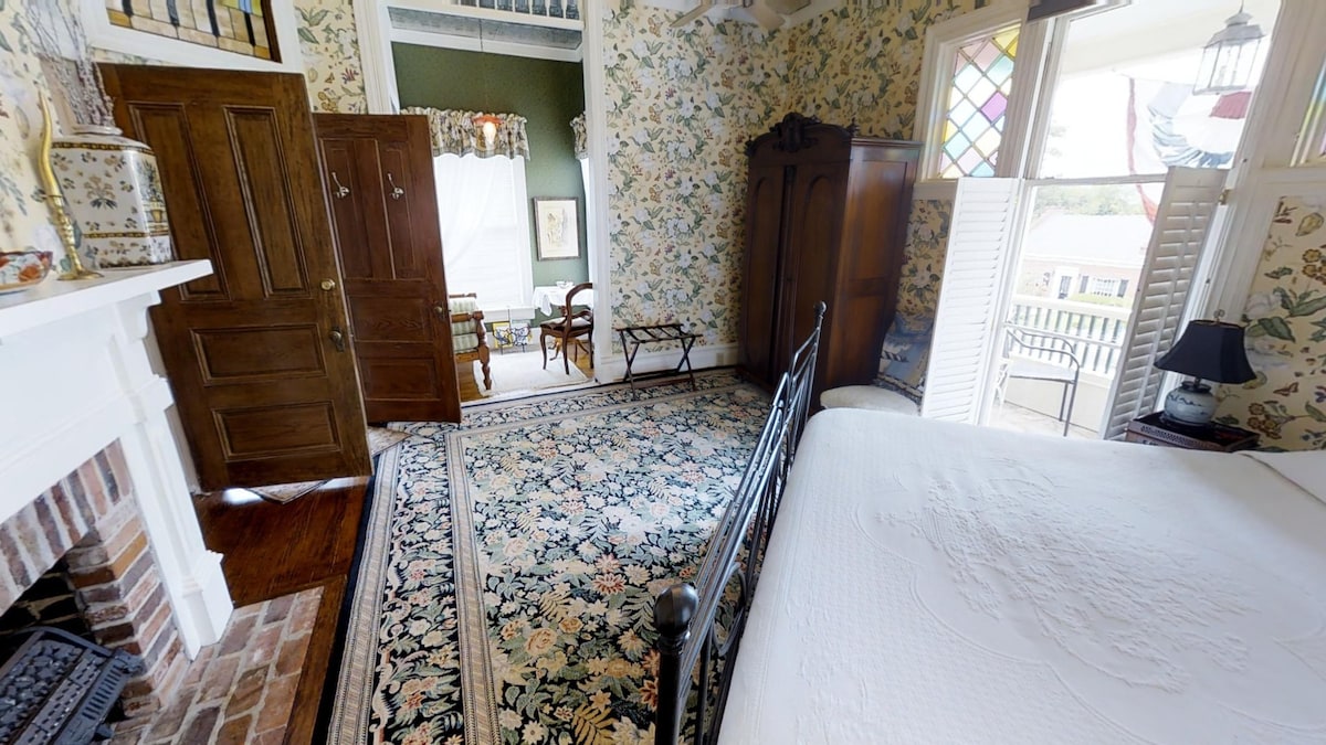 The room features floral wallpaper and a patterned rug, creating a cozy ambience. A black metal bed frame complements the space. A wardrobe and fireplace are visible, while a door leads to a balcony overlooking the street. A small dining area with a table and chairs is present.