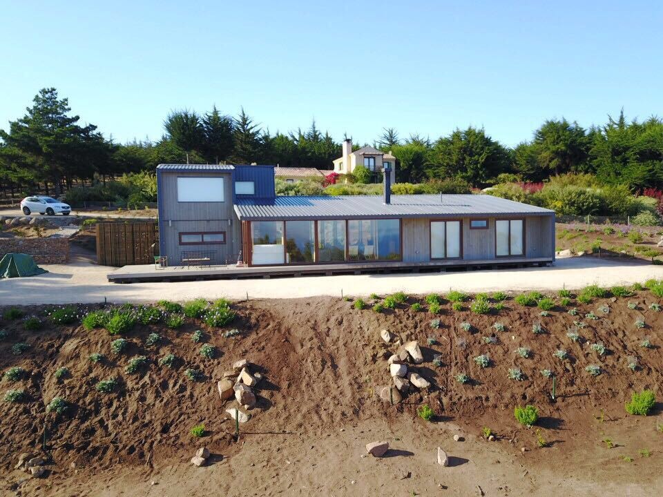 The exterior of a modern house is shown, featuring large windows and a sloped metal roof. The structure is surrounded by greenery, with a dirt path leading to the entrance, and a vehicle parked in the driveway nearby.