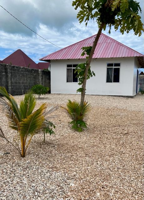 The exterior features a charming white house with a red roof, surrounded by a gravel yard. Small palm plants are positioned near the building, and the landscape is framed by a low wall and open sky, enhancing the tranquil setting.