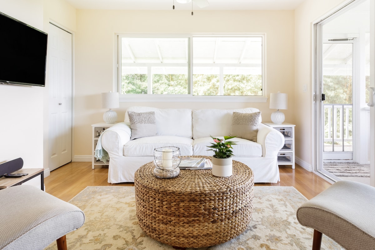 The living area features a soft white couch paired with light-colored accent chairs. A round wicker coffee table is positioned centrally, with a decorative plant placed on top. Large windows allow natural light to fill the space, connecting it to the outdoor porch.