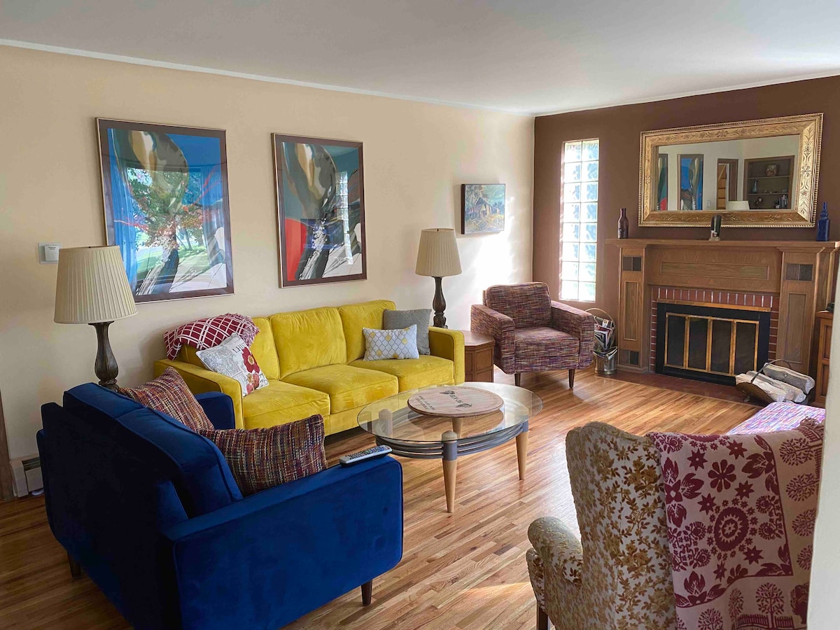 The living area showcases a blend of colorful seating options, including a bright yellow couch and a deep blue sofa. A coffee table sits centrally, surrounded by patterned armchairs. Artwork adorns the walls, complemented by natural light filtering through a glass block window, illuminating the wooden flooring.