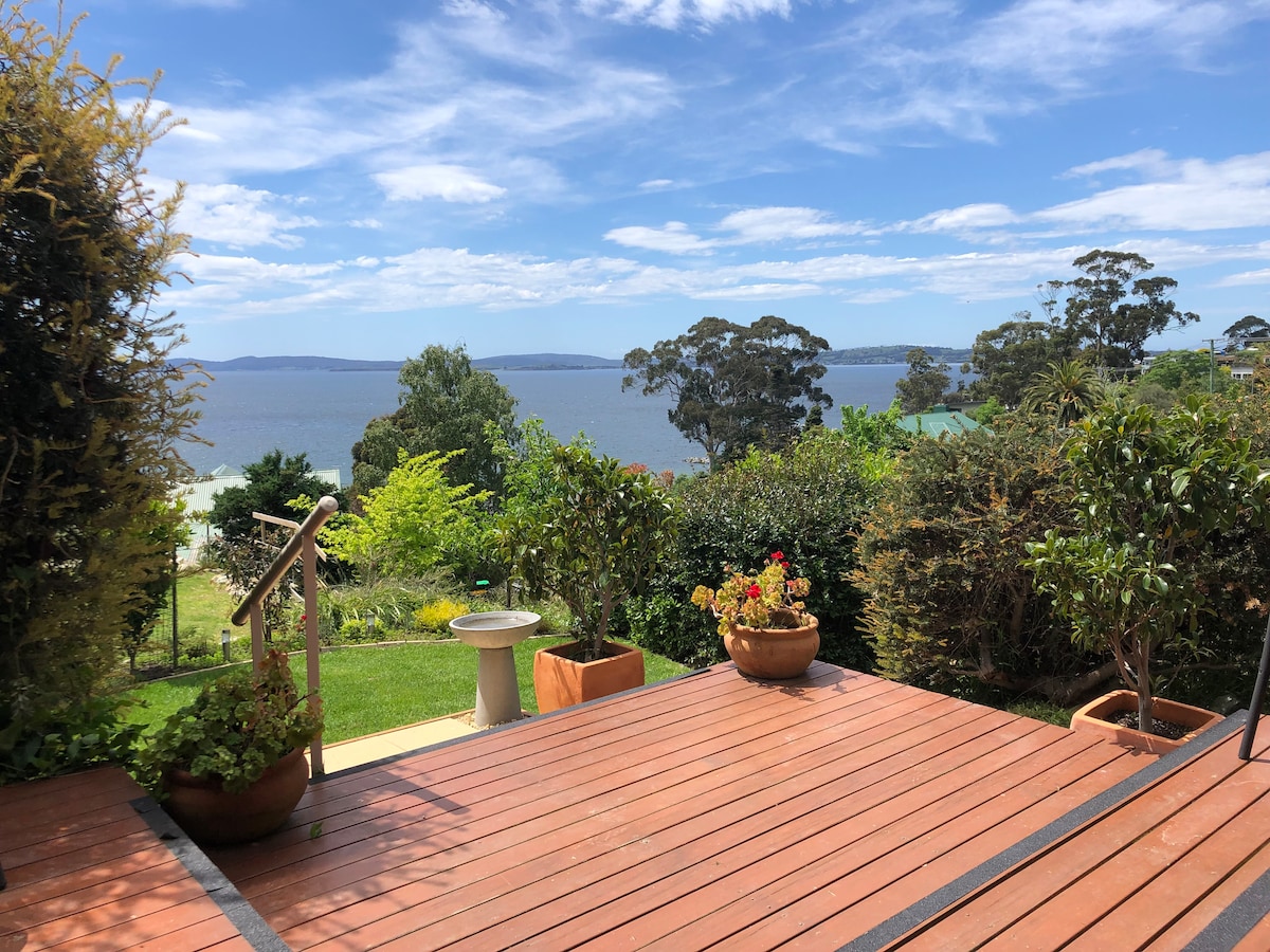 The private deck features wooden flooring, bordered by lush greenery and potted plants. A clear view of Kingston Beach and the Derwent Estuary is visible, with the sky showcasing a mixture of clouds and blue. Steps lead down to the garden area.
