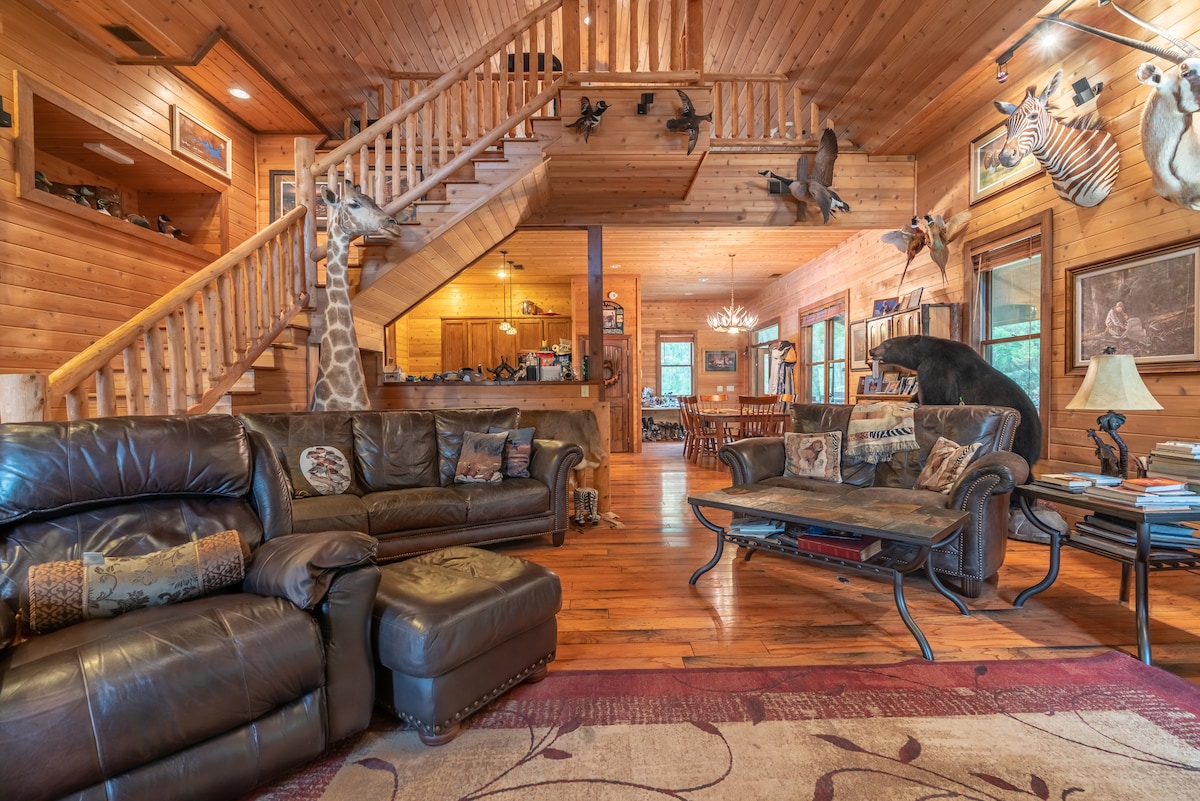 A spacious living area features leather seating arranged around a coffee table. The warm wooden interior is adorned with animal mounts and artwork. A staircase leads to an upper level, while an open area in the back includes a kitchen and dining space, enhancing the communal atmosphere.