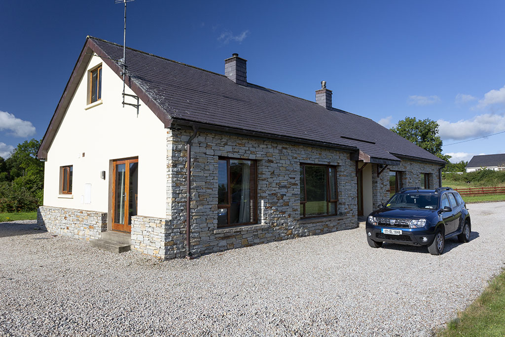 A modern guesthouse is featured with a stone façade and large windows, allowing natural light to enhance its appearance. A gravel parking area is visible alongside the entrance, with a vehicle parked nearby. Lush greenery surrounds the building, contributing to a peaceful setting.