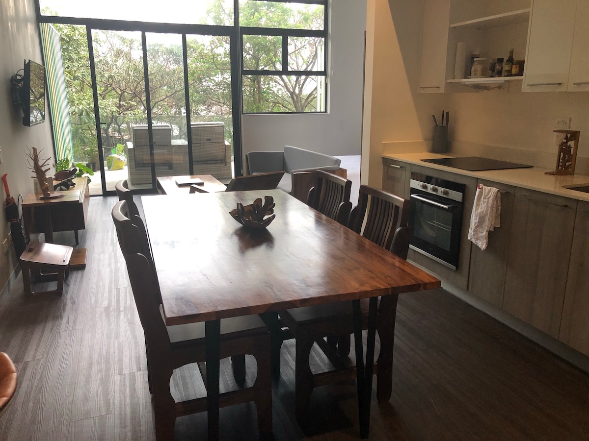 A spacious dining area is showcased with a large wooden table surrounded by eight chairs. A modern kitchen is visible in the background, featuring light cabinetry and an oven. Large windows allow natural light to fill the space, while greenery is seen through the glass.