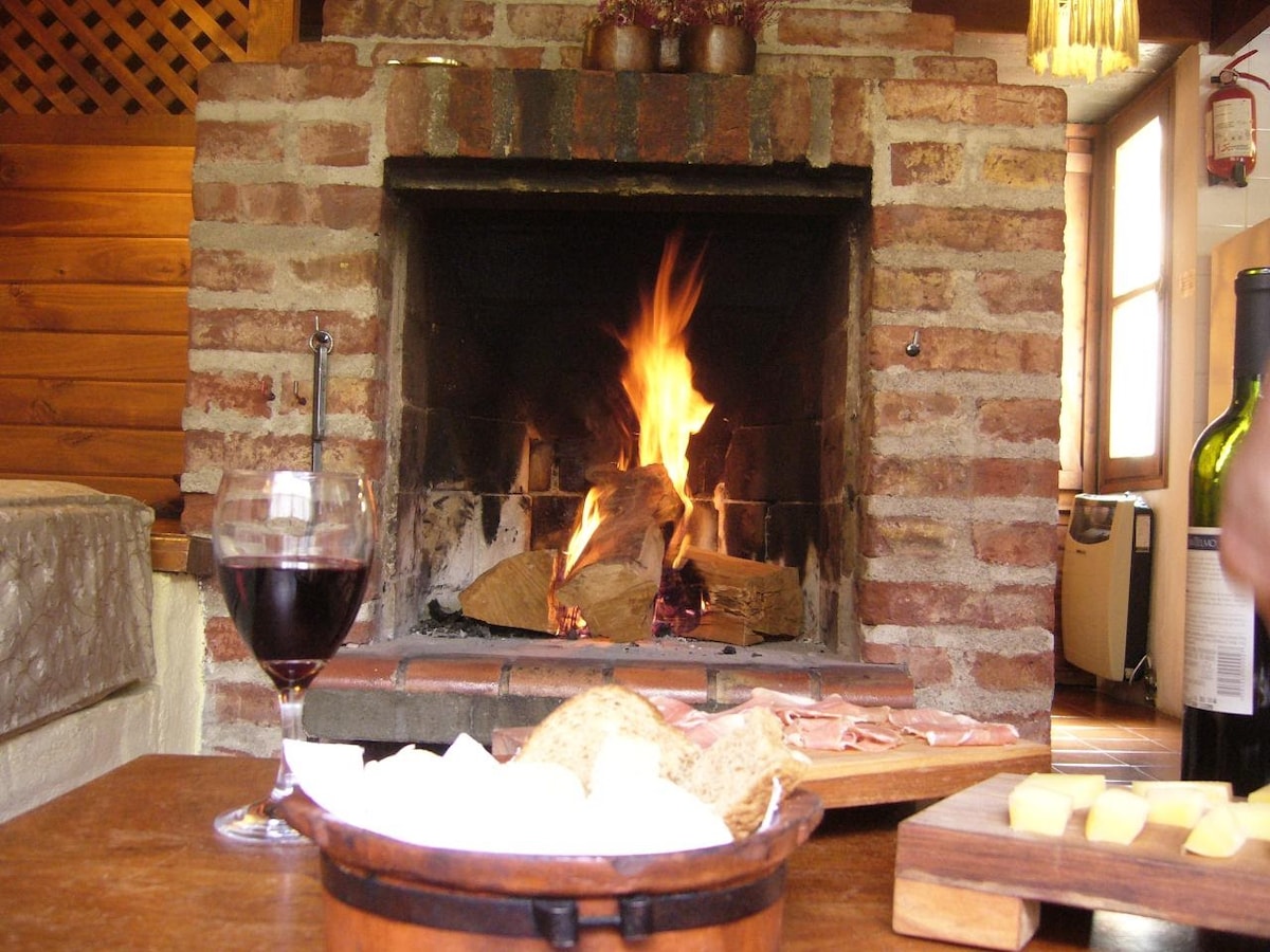 A cozy interior features a rustic brick fireplace with flames flickering warmly. A glass of red wine and a wooden bowl containing bread and cheeses are placed on a wooden table. The background showcases a wooden wall and additional furnishings, suggesting a welcoming atmosphere.