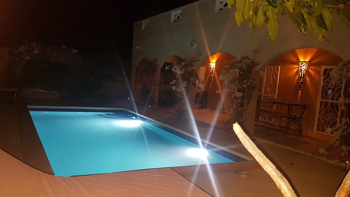 The nighttime view highlights a serene swimming pool, illuminated by underwater lights. Surrounding the pool, an outdoor seating area features elegant arches and subtle lighting, creating a calm and inviting atmosphere among lush landscaping.