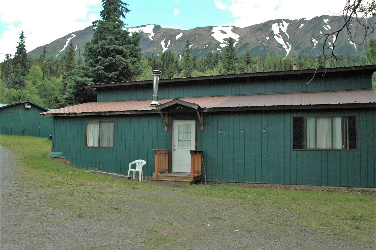 The exterior of a green cottage is displayed against a backdrop of snow-capped mountains. A wooden deck with steps leads to a white door at the center, flanked by large windows. A single white chair is positioned nearby on a gravel path.