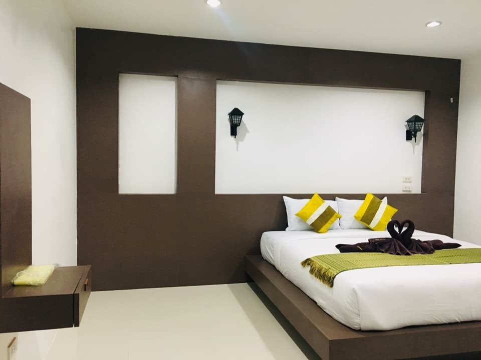 A modern bedroom features a spacious bed dressed in white linens, accented by vibrant yellow pillows. Wall sconces provide gentle lighting, while a sleek wooden shelf is mounted beside the bed. Neutral tones and clean lines contribute to a minimalist aesthetic.