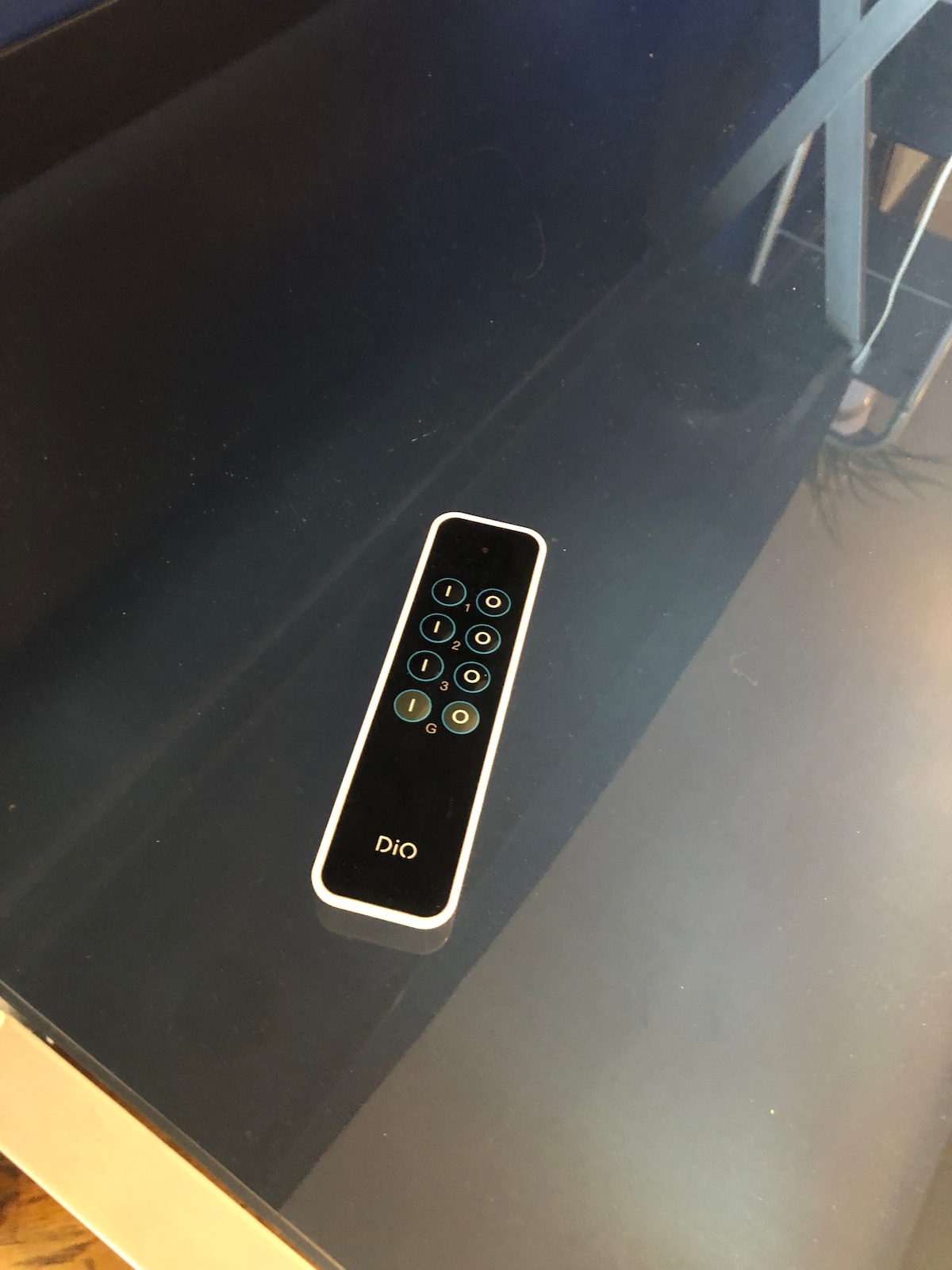 A sleek remote control rests on a reflective black table surface. The control features a minimalist design with several buttons, each marked with clear symbols. Soft lighting highlights the smooth edges of the remote and the table beneath it.